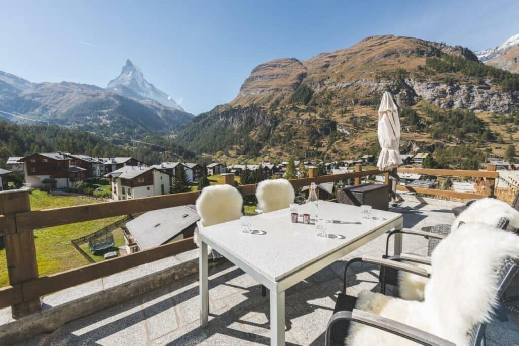 Varanda do Apartment Alpharmonie durante o dia com cadeira e quatro cadeiras do lado direito da imagem com vista para a cidade e montanhas. Representa onde ficar em Zermatt.