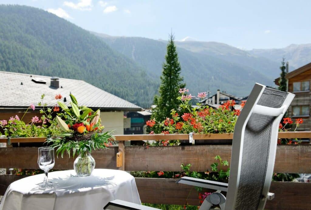 Imagem da varanda do Hotel Ambassador Zermatt com mesa redonda do lado esquerdo da imagem com duas taças e um vaso de flores em cima e uma cadeira do lado direito com vista para a cidade.