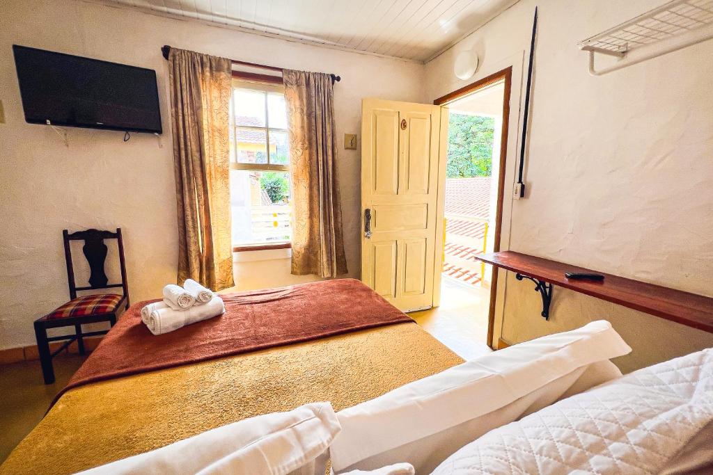 Quarto do Velinn Pousada Cesar. Uma cama de casal de frente para uma cadeira, uma televisão e a janela do quarto. Do lado direito a porta do quarto. Foto para ilustrar post sobre pousadas pet friendly em São Paulo.