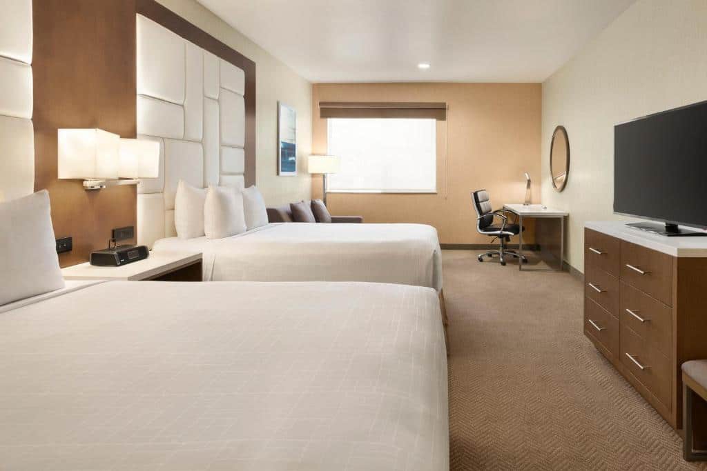 Quarto do Wyndham Anaheim. Duas camas de casal do lado esquerdo, entre elas uma cômoda com luminária. No fundo um sofá com abajur. Do lado direito uma cômoda, no fundo uma mesa de trabalho com espelho.