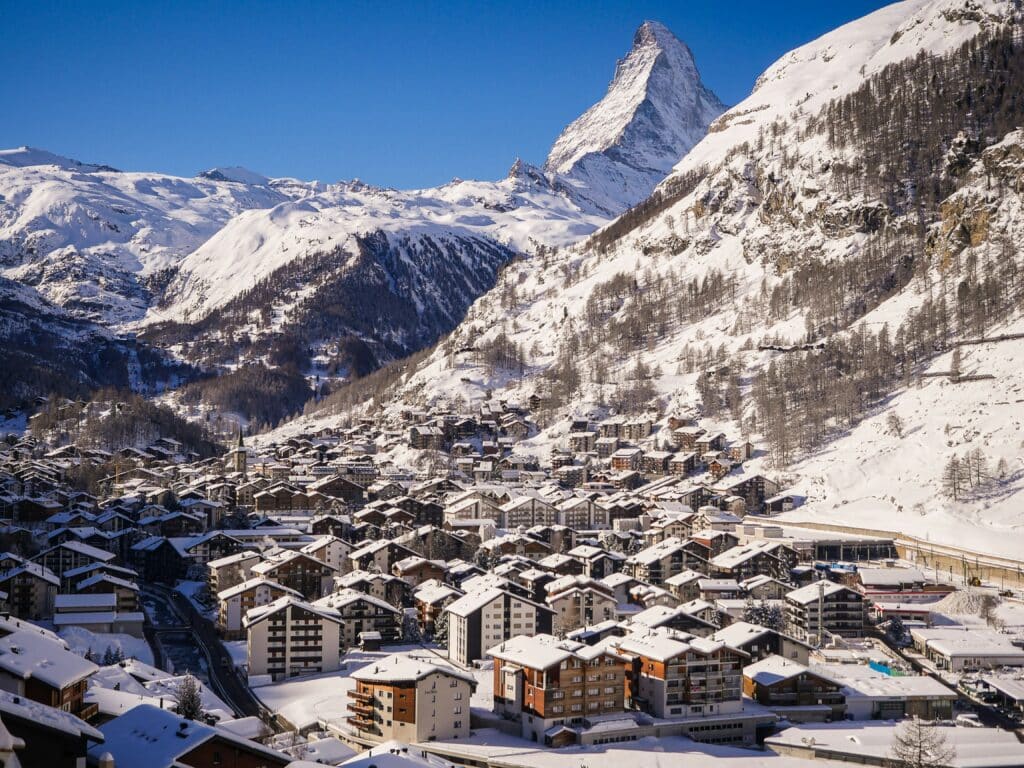 Imagem da cidade Zermatt durante o dia com várias casas a frente cobertas com neve e ao fundo montanhas com neve.