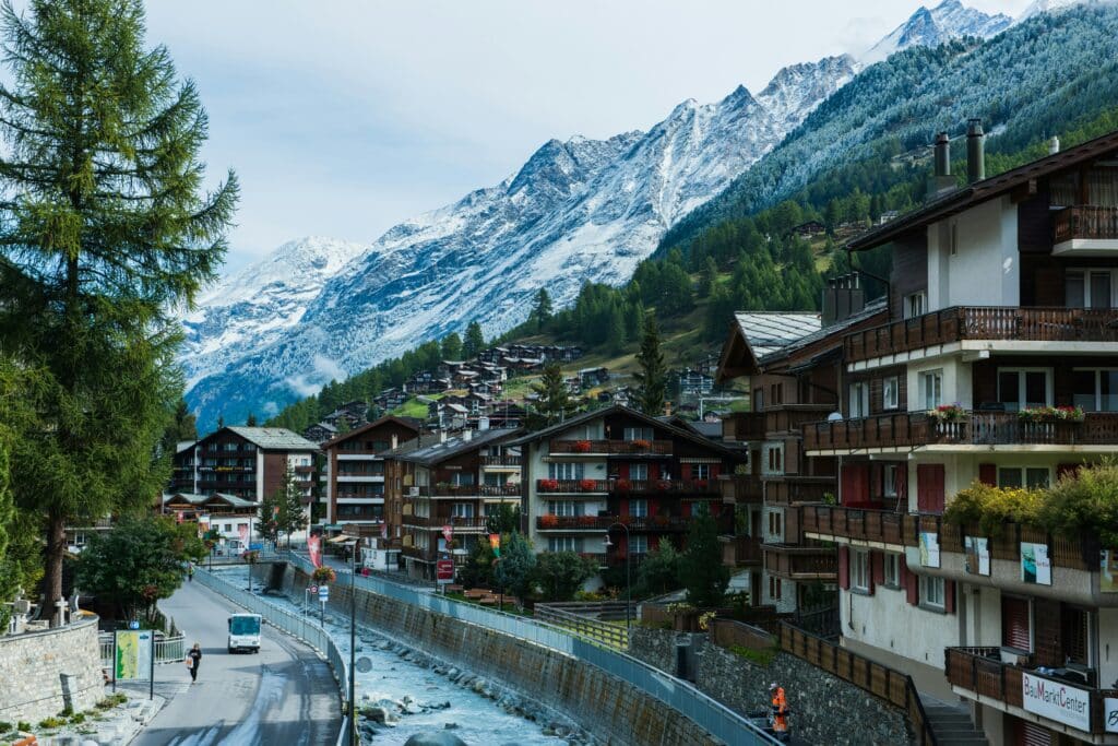 Imagem da cidade do Zermatt, Suíça durante o dia com um rio no centro e do lado direito vários prédios e ao fundo atrás dos prédios montanhas.