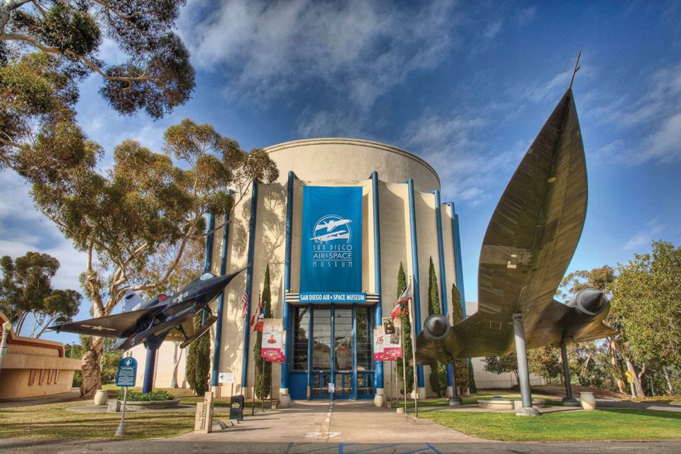 Entrada do San Diego Air & Space Museum. Ele tem uma estrutura grande, com representações de aviões e aeronavez decorando a parte da frente. Ao redor estão várias árvores.