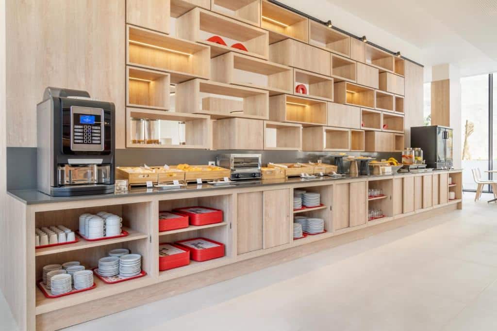 Área de refeições do Moov Hotel Lisboa Oriente com diversos itens de café da manhã, além de bandejas, pratos, canecas e máquinas expresso de bebidas quentes, para representar hotéis perto do Parque das Nações em Lisboa.