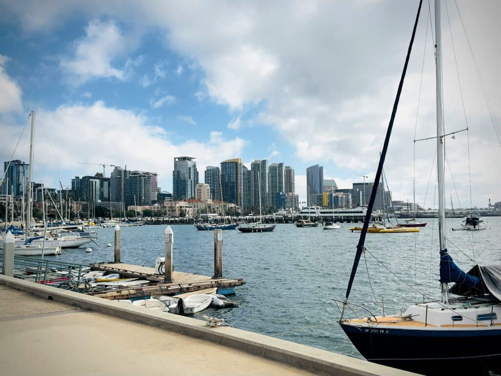 Foto de uma parte da Baiá de San Diego. Alguns barcos estão atracados em primeiro plano e outro estão soltos pela água. Ao fundo é possível ver também vários prédios da cidade.