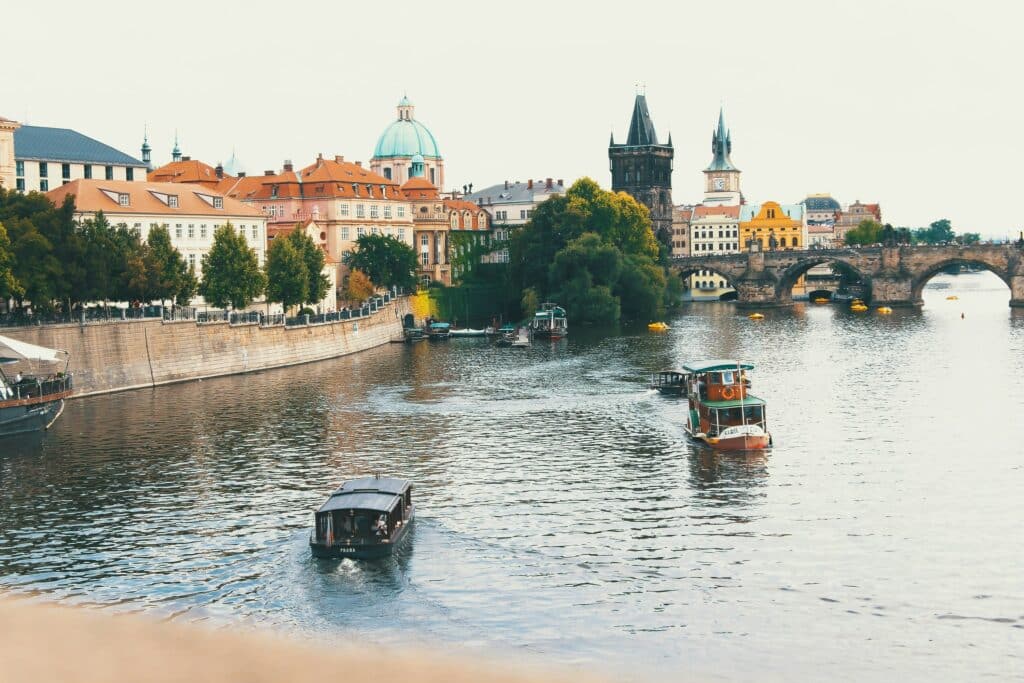 Imagem da cidade de Praga durante o dia com um rio a frente com dois barcos a frente ao fundo do lado direito a ponte e do lado esquerdo vários prédios históricos.