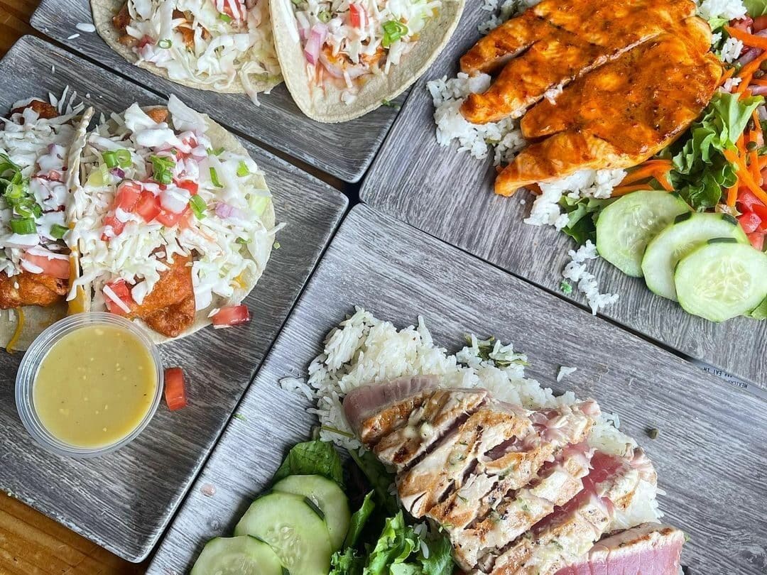 Foto de quatro pratos de comida no restaurante Blue Water Seafood Market and Grill. Nos pratos do lado esquerdo estão tacos recheados e molho. Do lado direito estão frutos do mar, arroz, legumes e verduras.