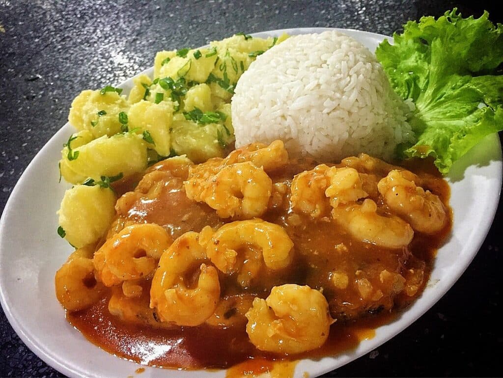 Um prato com camarão ao molho, arroz branco, salada de batata coberta com ervas e uma guarnição de alface ao lado. Esse é um prato da Cantina Montóia, em Águas de Lindóia.