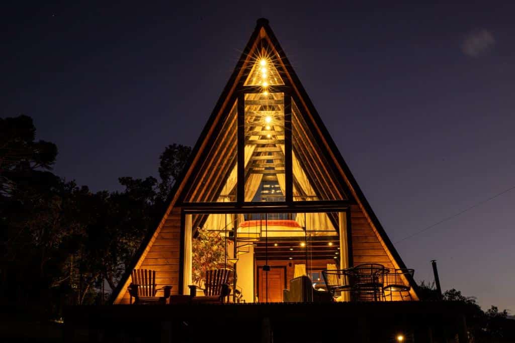 Chalé do Altitude 1140 Urubici - Chalés com fachada de vidro em formato triangular. Na frente do chalé tem algumas cadeiras, e por dentro do chalé a iluminação é amarelada. Em volta tem algumas árvores durante o anoitecer. Imagem ilustrando post Chalés em Urubici.