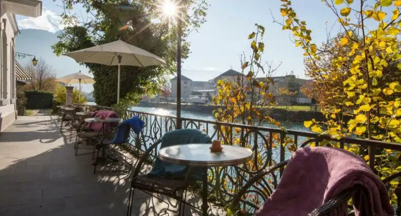 Imagem da varanda do Boutique Hotel Bellevue durante o dia com cadeiras e mesas no ambiente, o local oferece vista para o lago e a cidade, para representar hotéis em Interlaken.