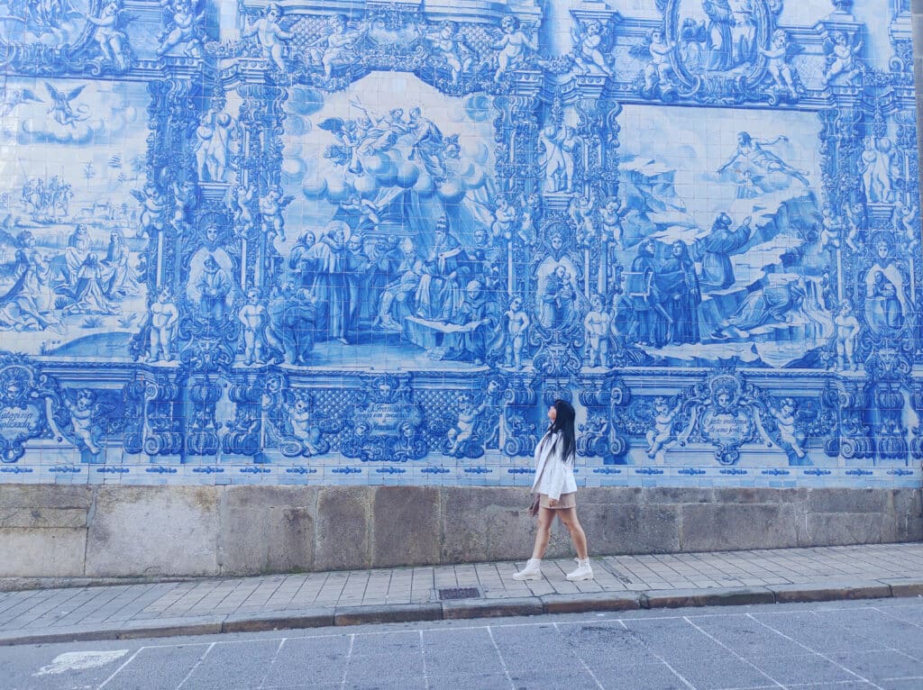 Uma pessoa vestida de branco está em frente a um grande mural azul e branco em uma parede de azulejos azuis, a Capela das Almas, em Porto, Portugal, olhando para a obra de arte.