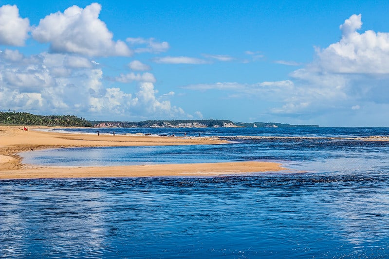 Foto de uma praia em Caraíva, na Bahia. Mostra um mar bem azul, e parte da areia, na direita. O céu está azul e com algumas nuvens.