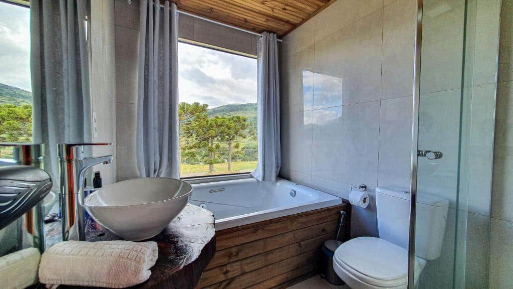 Banheiro do Cascata dos Amores com banheira abaixo de uma janela com vista para a natureza. Há ainda pia e vaso, todas as louças em branco e detalhes em madeira.