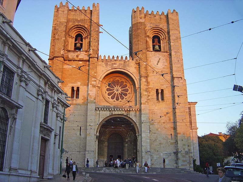 Imagem da Catedral da Sé durante o dia com edifício do lado direito no meio uma rua e a frente a igreja. Representa o que fazer em Lisboa.
