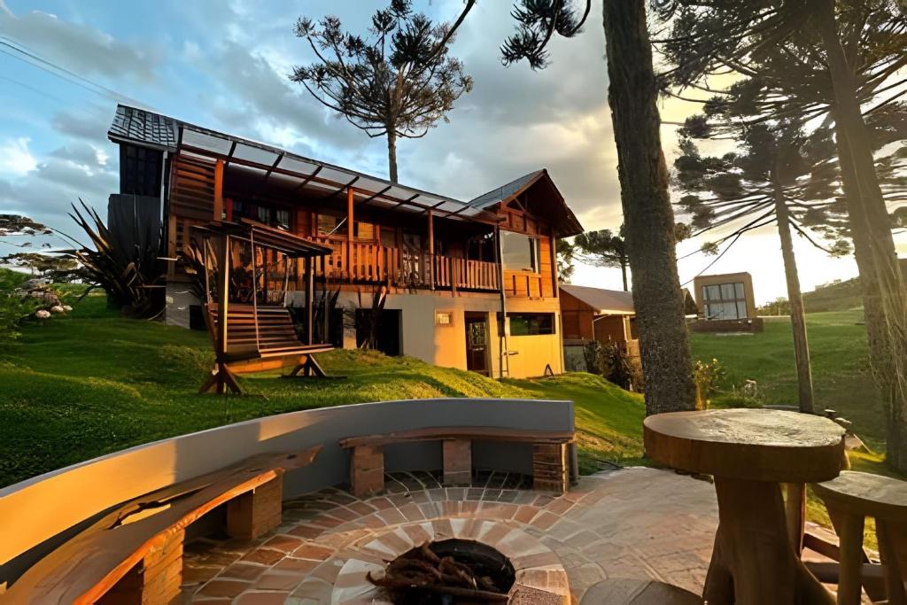 Área exterior do Chalé Urubici - Morada nas Montanhas mostrando a fachada em dois andares da estadia feita da madeira com um banco semicircular onde é possível observar a natureza. Há araucárias ao redor.