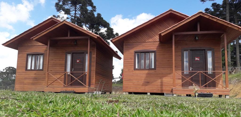 Fachada do Chalés Sossego da Serra sendo casinhas de madeira com porta e janela de vidro, há uma grama baixa bem cuidada abaixo, o céu é azul com nuvens e há araucárias atrás.