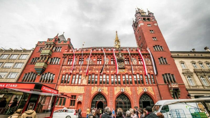 Imagem do edifício da Prefeitura da Basiléia durante o dia com pessoas a frente e ao fundo o prédio.