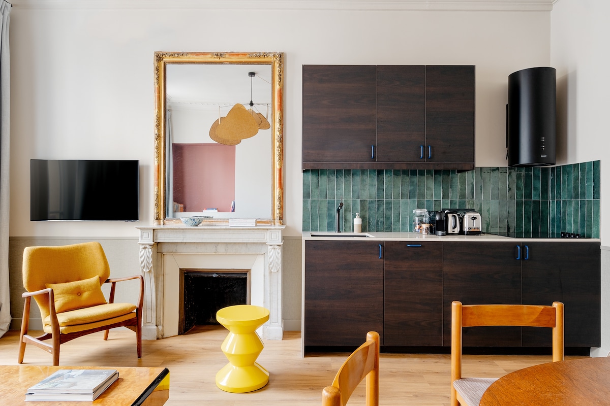 Sala de estar de uma das Edgar Suites Saint-Lazare - Amsterdam. Ao lado direito ficam as bancadas e armários com utensílios de cozinha, e ao lado está uma TV na parede atrás de uma poltrona.