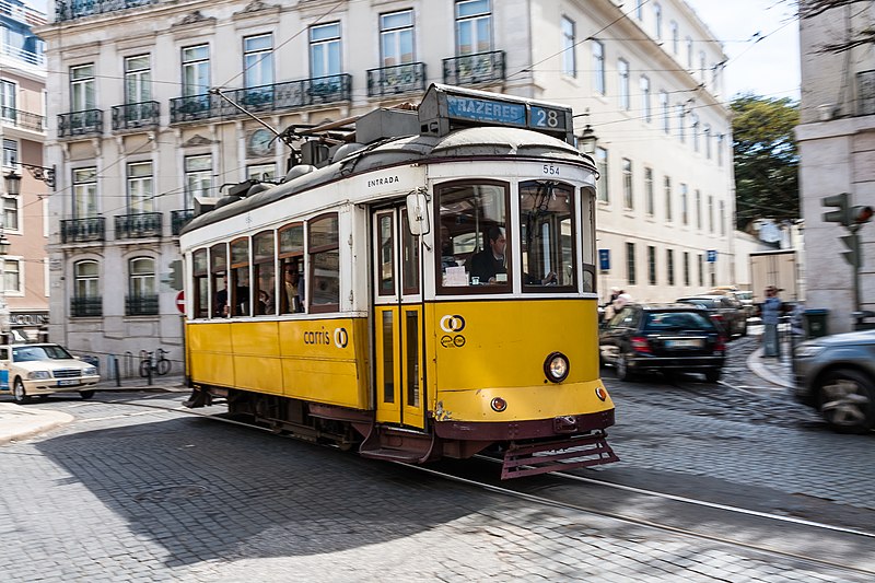 Imagem de um Eléctrico 28 durante o dia com o bondinho no centro da imagem andando no meio da duas do lado direto carros e ao fundo prédios. Representa o que fazer em Lisboa.