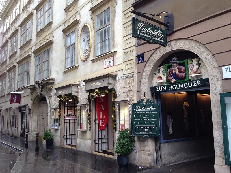 Cena de rua mostrando uma joalheria com uma grande placa de "VENDA" e o restaurante Zum Figlmüller com duas entradas proeminentes.