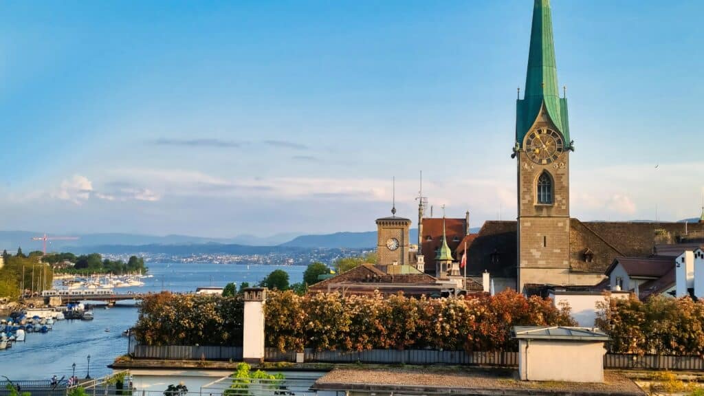 Imagem da Igreja de Fraumünster do lado direito da imagem durante o dia com casas em volta e do lado esquerdo o rio.
