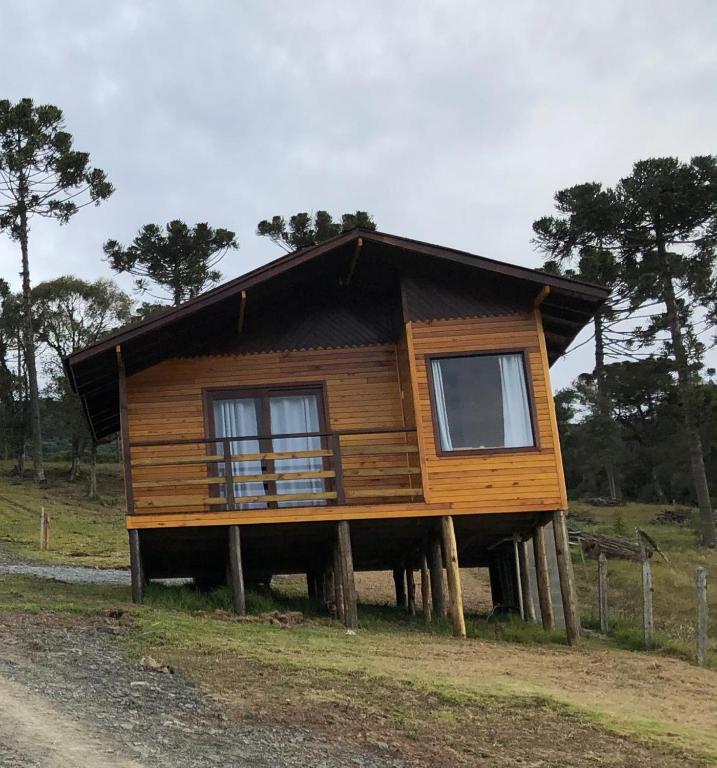 Um dos chalés em Urubici na Hospedagem Família Cruz sendo uma casinha no morro totalmente em madeira, com janela e porta de vidro com cortinas e uma varanda. A grama abaixo é baixa e há araucárias ao fundo.