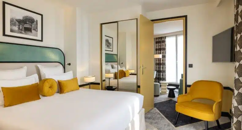 Quarto do Hotel Aiglon, um dos hotéis em Montparnasse. Uma cama de casal está encostada na parede ao lado esquerdo com mesinhas de cabeceira e abajures dos dois lados, assim como um quadro na parede TV e uma poltrona logo em frente, e na lateral direita fica um guarda-roupa de portas espelhadas. Uma porta ao lado do armário leva à sala de estar, onde é possível ver sofás, mesinhas e uma janela com cortinas.