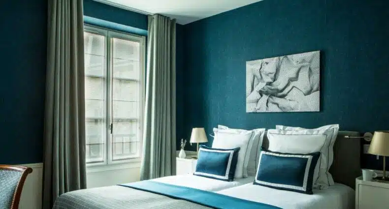 Quarto do Hôtel de la Place du Louvre - Esprit de France, um dos hotéis bem localizados em Paris. A cama de casal está encostada na parede central com mesinhas de cabeceira e abajures dos dois lados, assim como um quadro na parede acima. Ao lado esquerdo fica uma janela com cortinas.