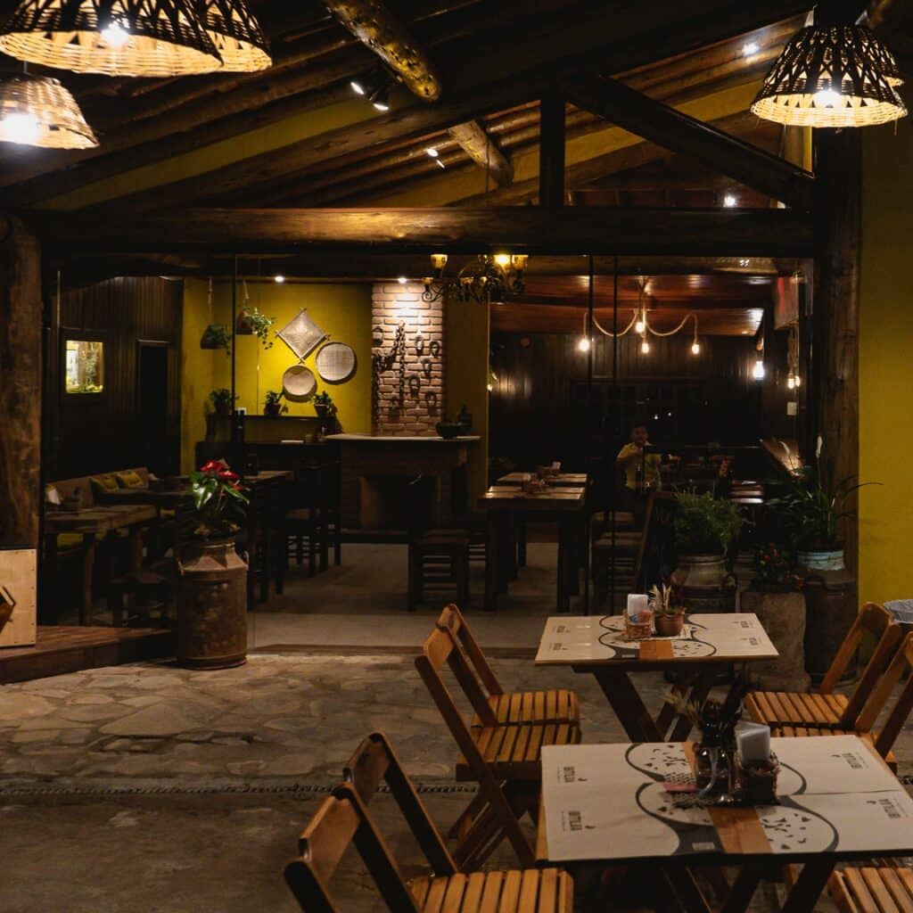Restaurante Ibitilua que mostra duas mesas com cadeiras de madeira na parte de fora e do lado de dentro do restaurante tem mais mesas. A decoração é rústica com luzes e plantas espalhadas.