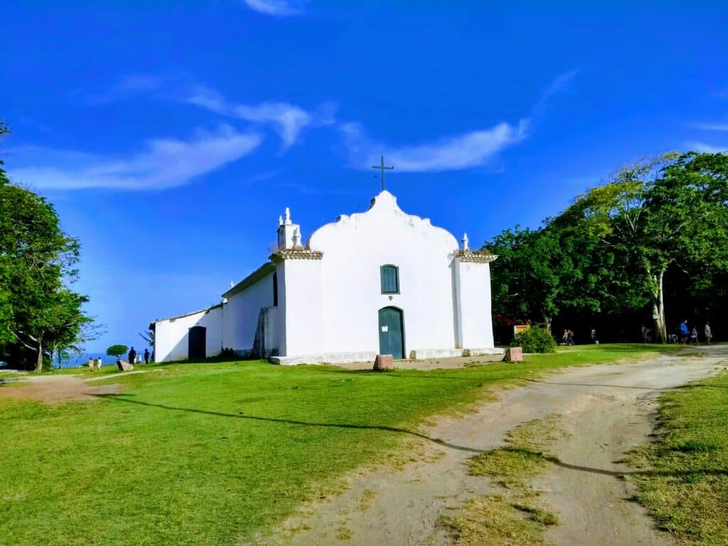 Foto da Igreja de São João Batista, em Trancoso. Está sobre um gramado bem verde, com árvores ao fundo dos dois lados. O céu atrás é bem azul e com poucas nuvens. Há algumas pessoas ao lado esquerdo da igreja, bem no fundo da foto.