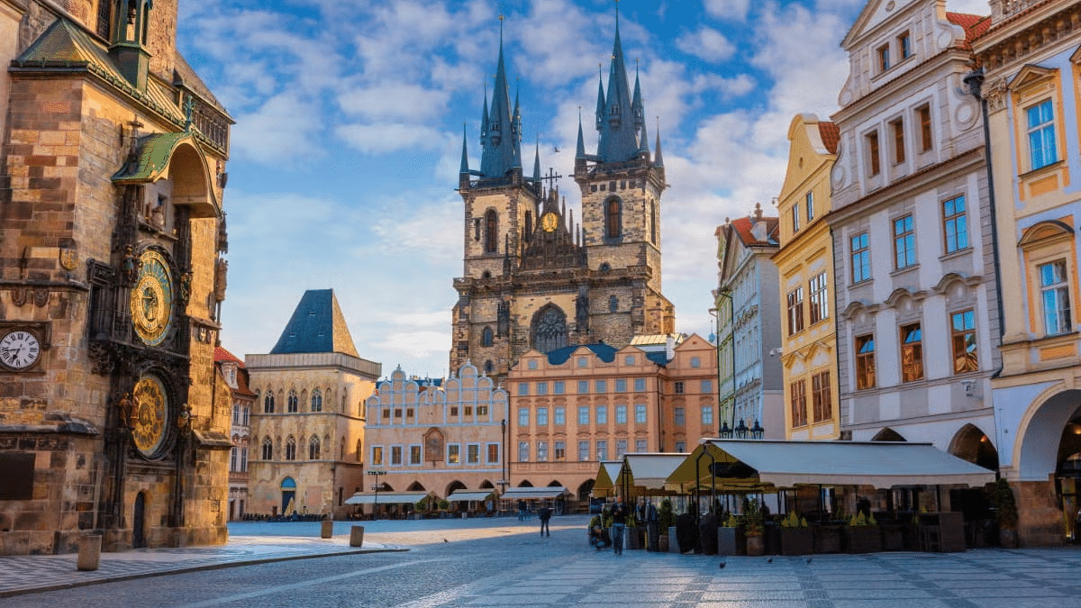 Imagem da cidade de Praga durante o dia com um pátio a frente ao fundo prédios históricos e mais ao fundo uma igreja. Representa o que fazer em Praga.
