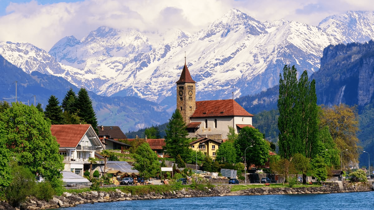 Imagem durante o dia de um rio a frente e ao fundo árvores e alguns edifícios.