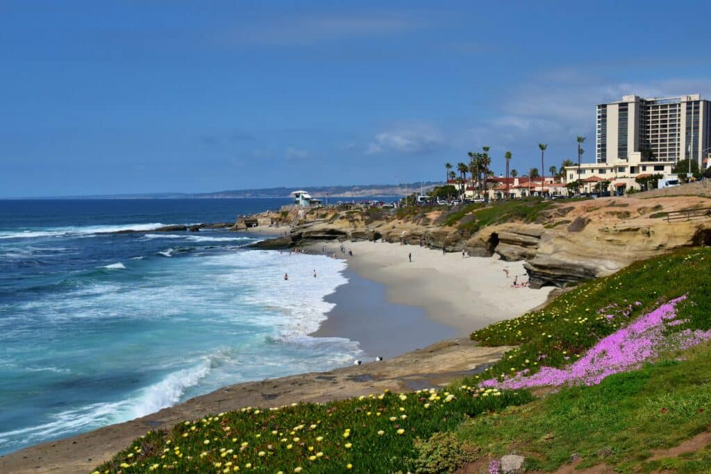 Praia de La Jolla Cove. O mar está do lado esquerdo e a faixa de areia e pedras que cercam o ambiente estão do lado direito. Ao fundo é possível ver também vários prédios e casas.