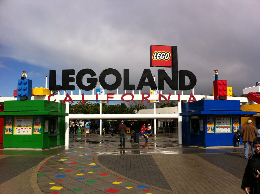 Entrada do Parque Legoland Califórnia. Há um letreiro bem no meio e vários legos decorando o local. Algumas pessoas andam por ali.