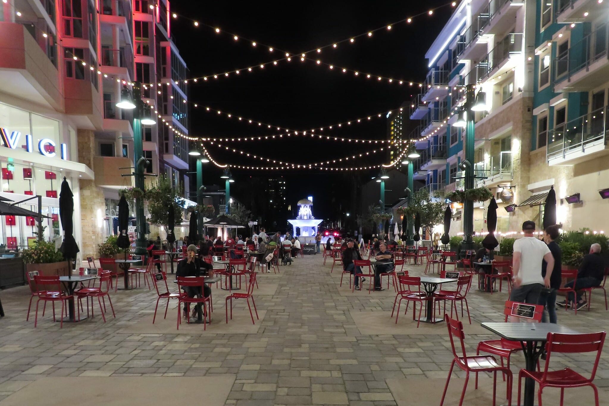 Piazza della Famiglia em Little Italy, praça em que uma fonte de água toda iluminada está no final. Antes dela estão diversas mesas e alguma lojas.