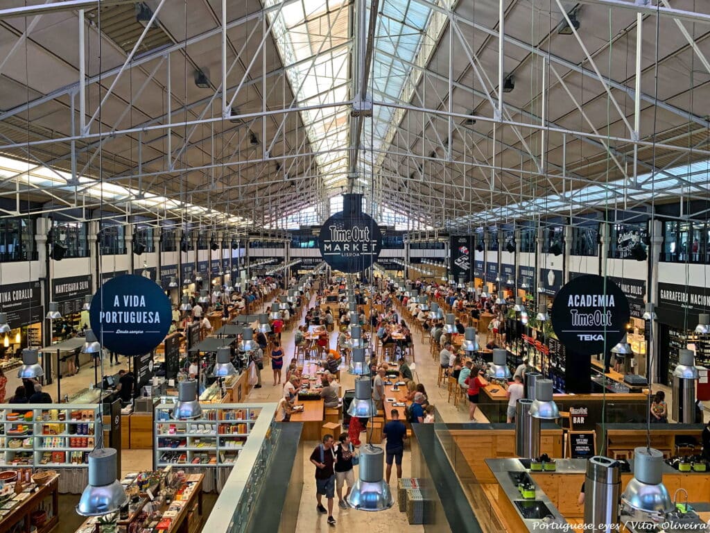 Imagem de dentro do Mercado da Ribeira vista de cima com pessoas e mesas abaixo compondo o ambiente.
