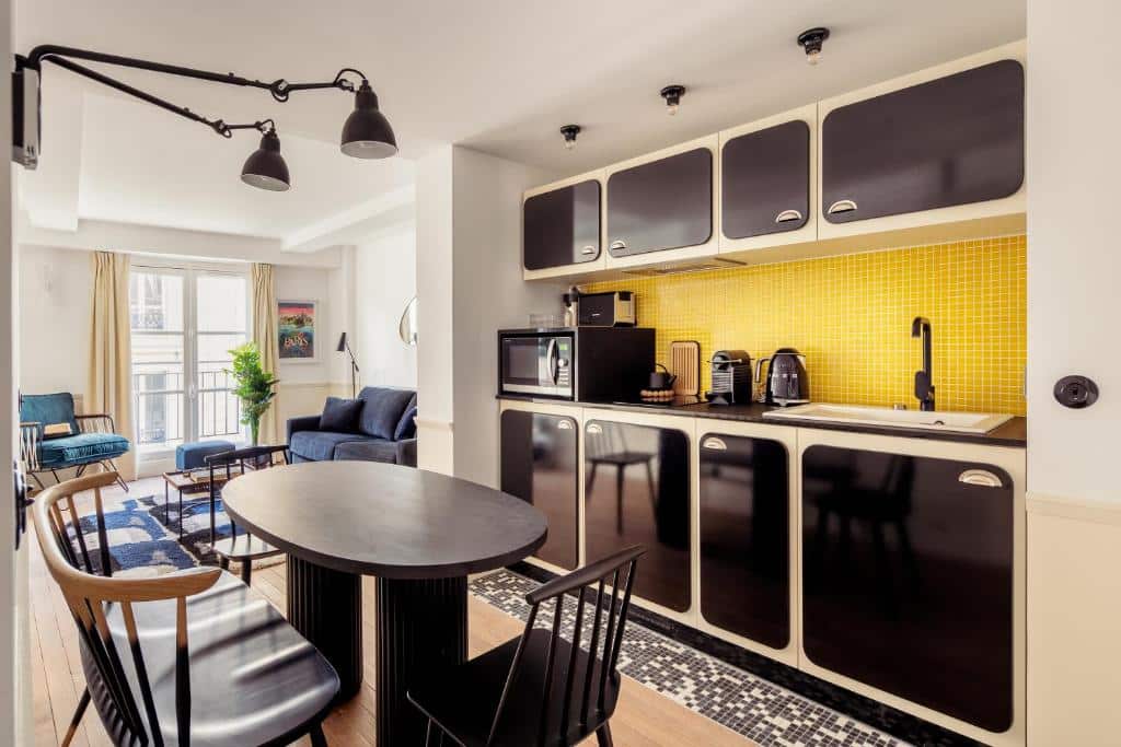 Cozinha da My Maison In Paris - Louvre, onde uma mesa com banquinhos encara armários e utensílios na parede ao lado direito. Há um micro-ondas, uma chaleira e uma cafeteira ao lado da pia. Ao fundo do cômodo fica a sala de estar com sofá, poltronas, TV e uma janela com cortinas.