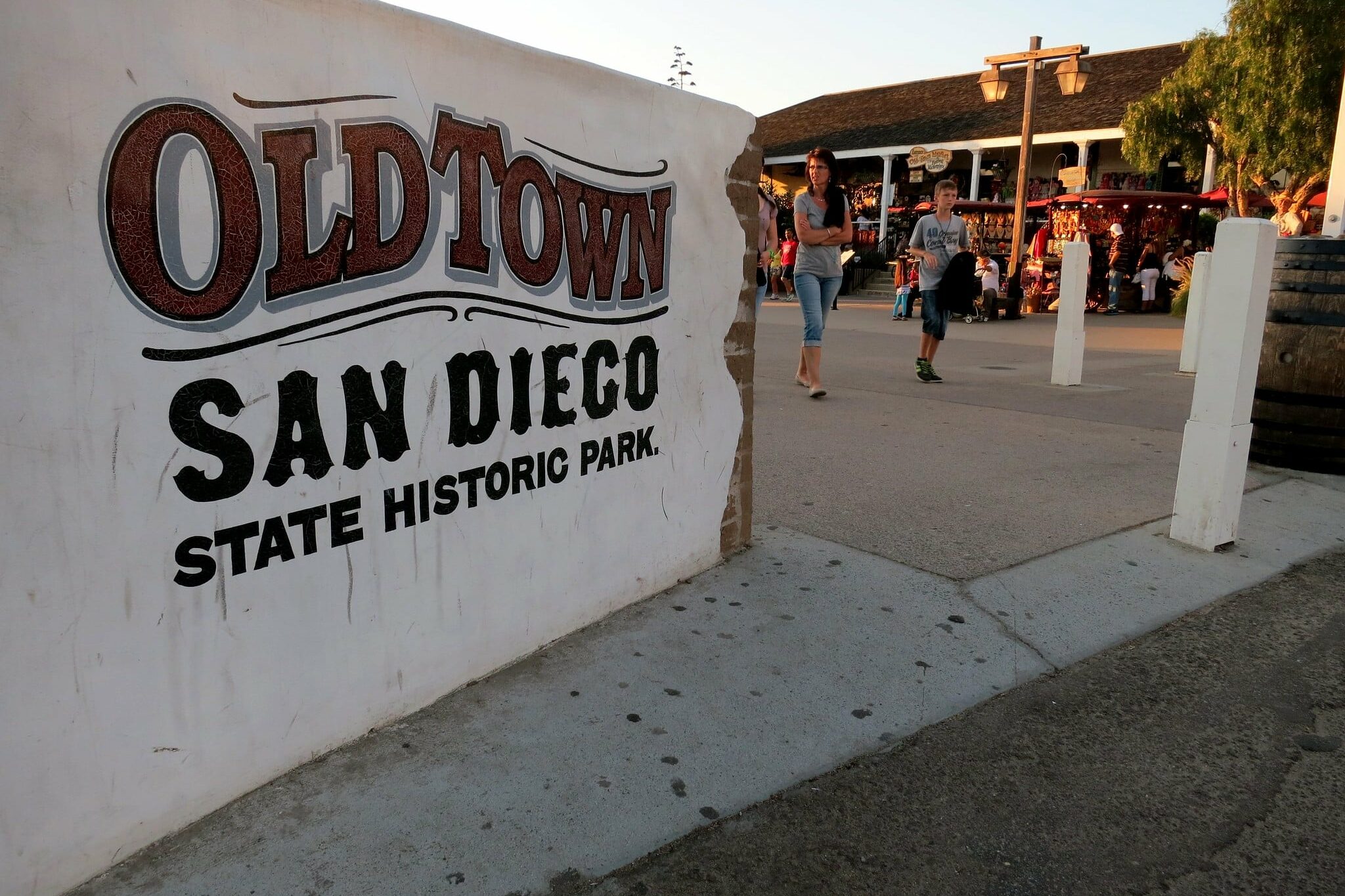 Parede escrito "Old Town San Diego State Historic Park" indicando a entrada de Old Town. Ao fundo é possível ver algumas barracas e pessoas andando. A foto está na lista de o que fazer em San Diego.