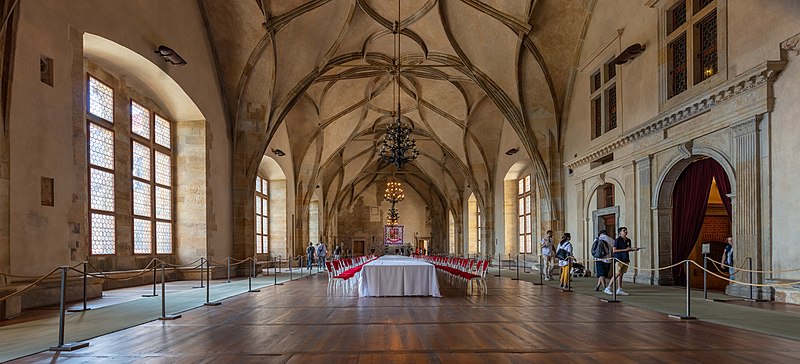 Imagem do interior do Palácio Real no centro da imagem ao fundo uma mesa quadrada comprida com várias cadeiras do lado direito pessoas andando. Representa o que fazer em Praga.