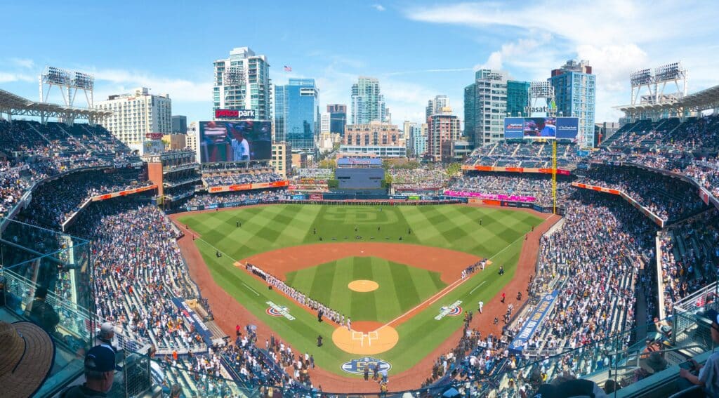 Vista panorâmica do estádio PETCO Park. O campo está ao meio e ao redor toda a arquibancada cheia. Os telões mostram jogadores e ao fundo, como o estádio é aberto, está a cidade de San Diego.