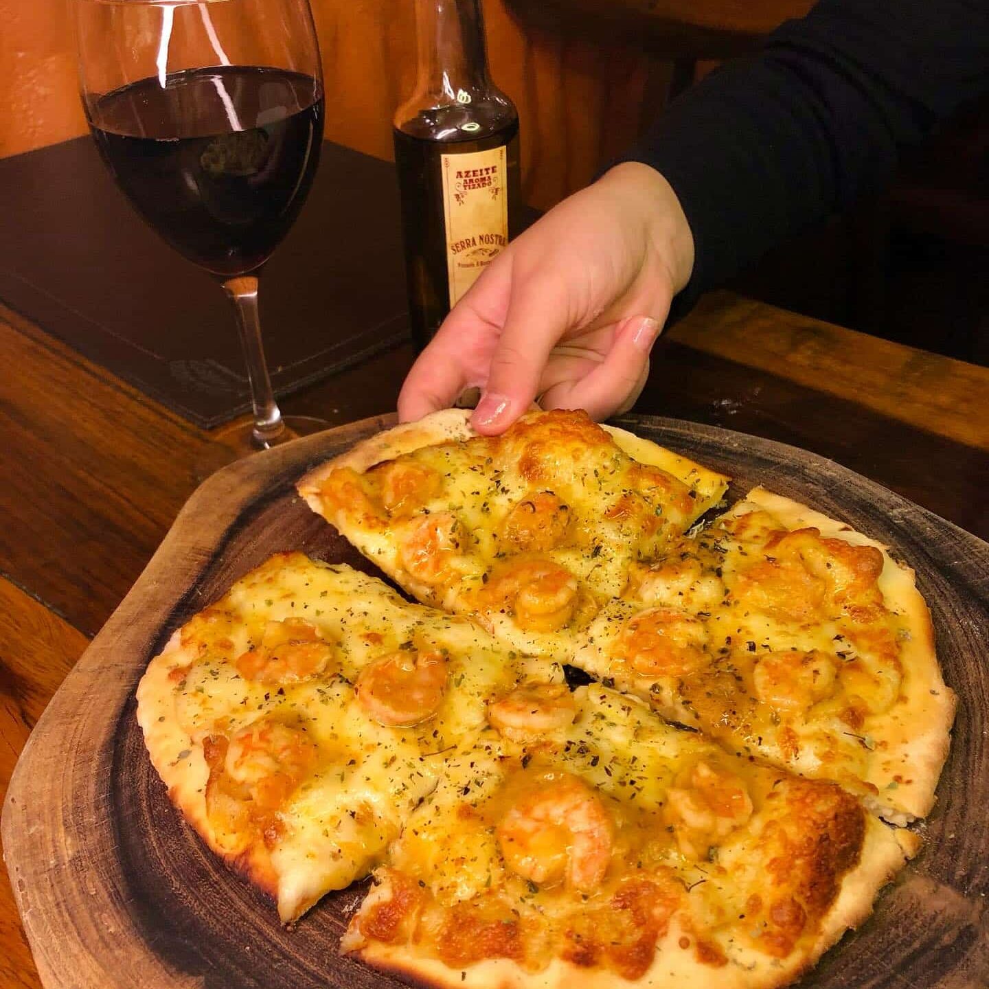 Pizza de camarão da Pizzaria Serra Nostra com uma mão segurando um pedaço e ao fundo uma taça de vinho com uma garrafa de azeite ao lado. Imagem ilustrando post O que fazer em Ibitipoca.