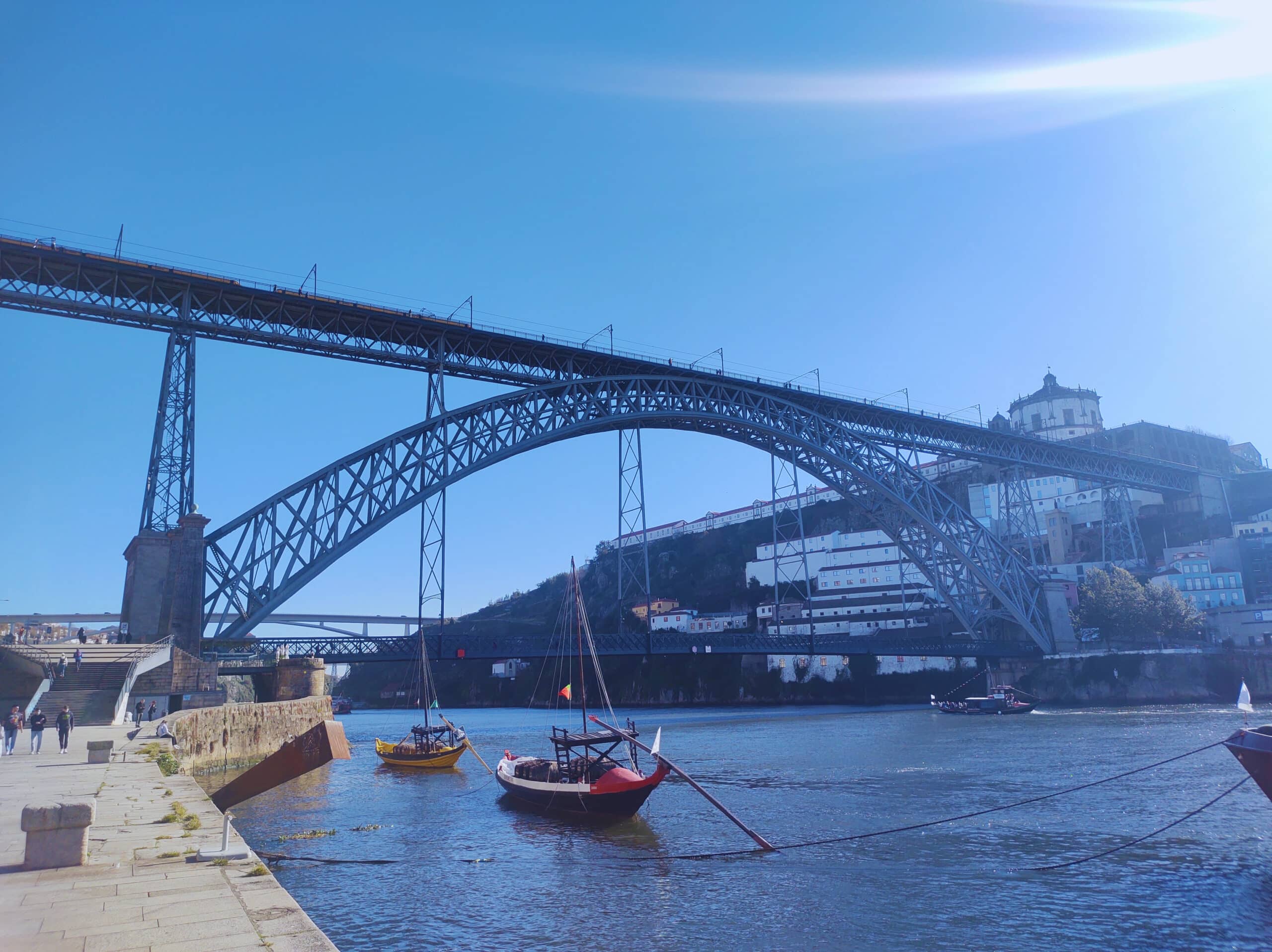 Ponte D. Luís I durante o dia com barcos do lado direito da imagem dentro do rio e ao fundo a ponte. Representa roteiro para Portugal.