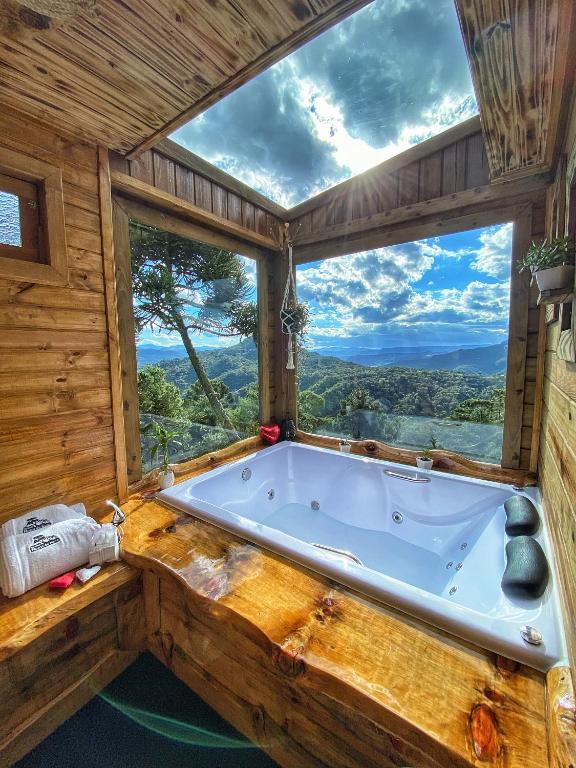 Banheira grande na Pousada Flocos de Neve com janelas abertas de ambos os lados em que é possível ver a natureza e o céu azul. O interior é de madeira, e há locais para toalhas de banho.