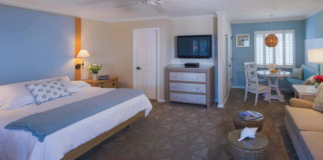 Quarto do Scripps Inn. Do lado direito um sofá, duas mesinhas de centro, no fundo uma mesa de jantar e janela. Do lado esquerdo uma cama de casal, duas cômodas e um abajur, uma TV e uma porta.