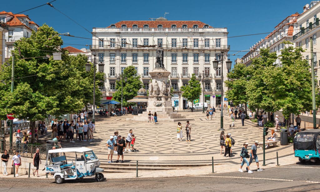 Imagem da Praça Luis de Camões durante o dia com pessoas andando no pátio a frente e em volta prédios.