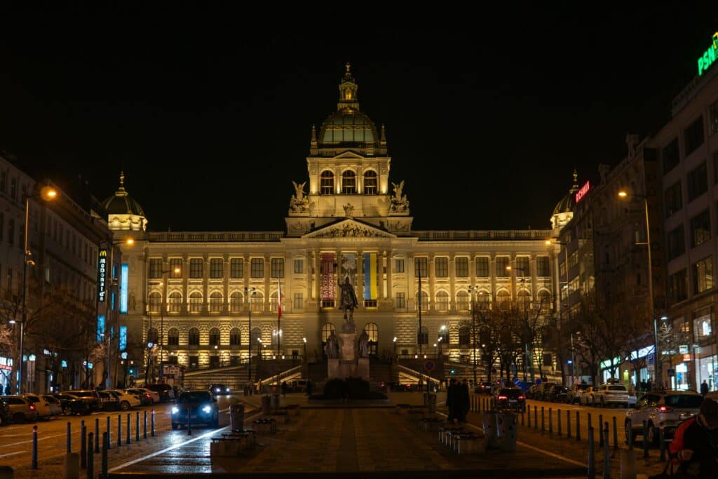 Imagem de noite da Praça Venceslau com um pátio a frente e do lado direito e esquerdo ruas com veículos e ao fundo um prédio grande. Representa o que fazer em Praga.