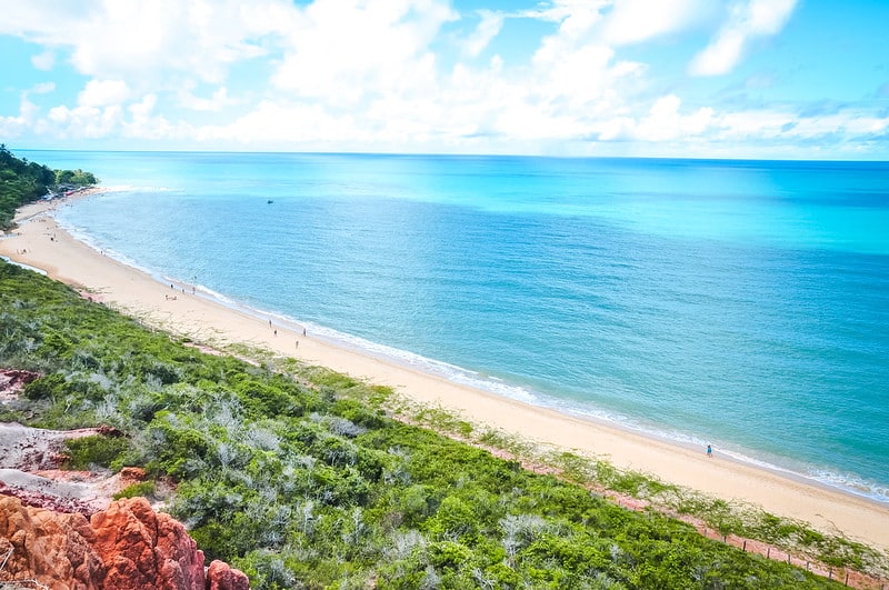 Praia de Arraial d'Ajuda. na Bahia. É uma foto tirada no topo de algum morro ou colina, ou até mesmo aérea, que mostra o um mar azul e cristalino. No canto inferior direito vemos uma colina coberta por bastante verde, e lá embaixo está a areia e a praia. Algumas pessoas caminham pela areia.