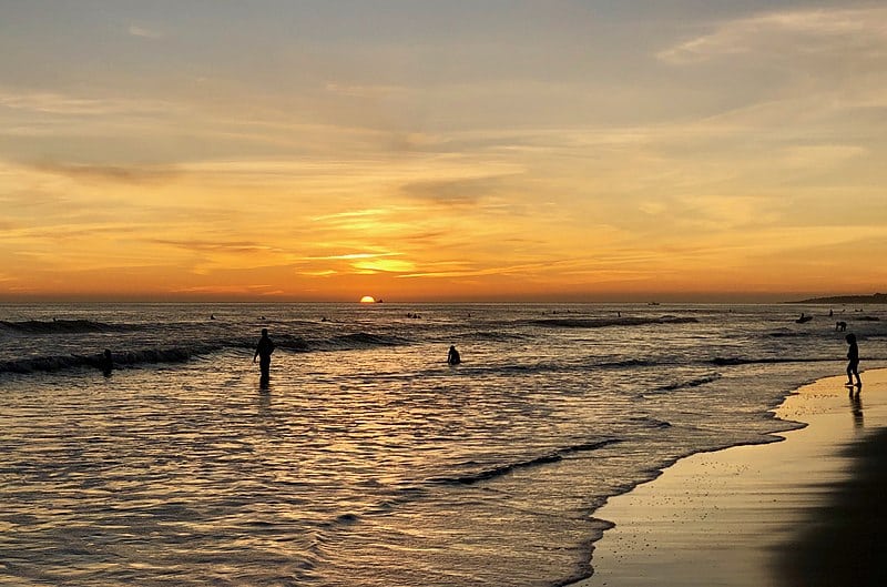 Imagem da Praia de Carcavelos durante o final do dia com faixa de areia do lado direito no canto e do lado esquerdo o mar com algumas pessoas dentro. Representa o que fazer em Lisboa.
