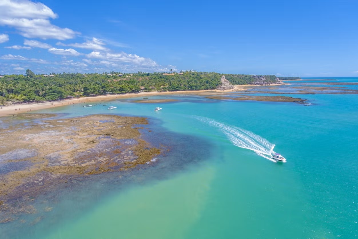 Uma foto aérea e panorâmica da Praia do Espelho, entre Trancoso e Caraíva. Mostra a água azul cristalina, com uma lancha passando, além do resto da geografia que compõe o lugar. Há árvores ao fundo. O céu é azul e com poucas nuvens.