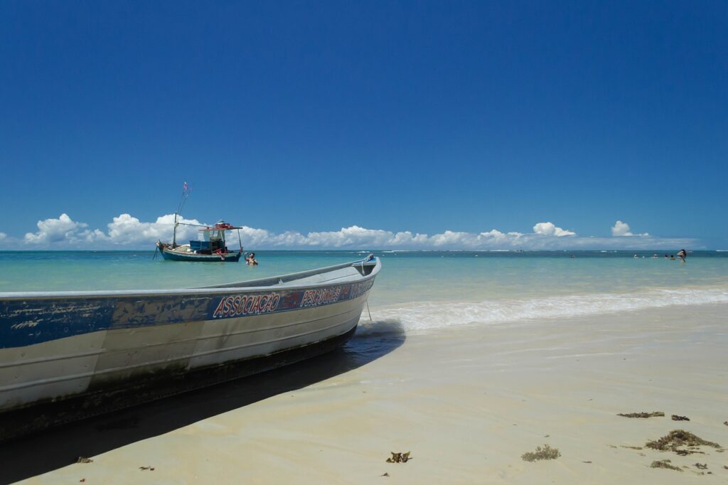 Foto da Praia dos Coqueiros, em Trancoso. Há uma canoa na areia, perto do mar. No mar, ao fundo, há um barco. O céu é azul e com algumas nuvens perto do horizonte.